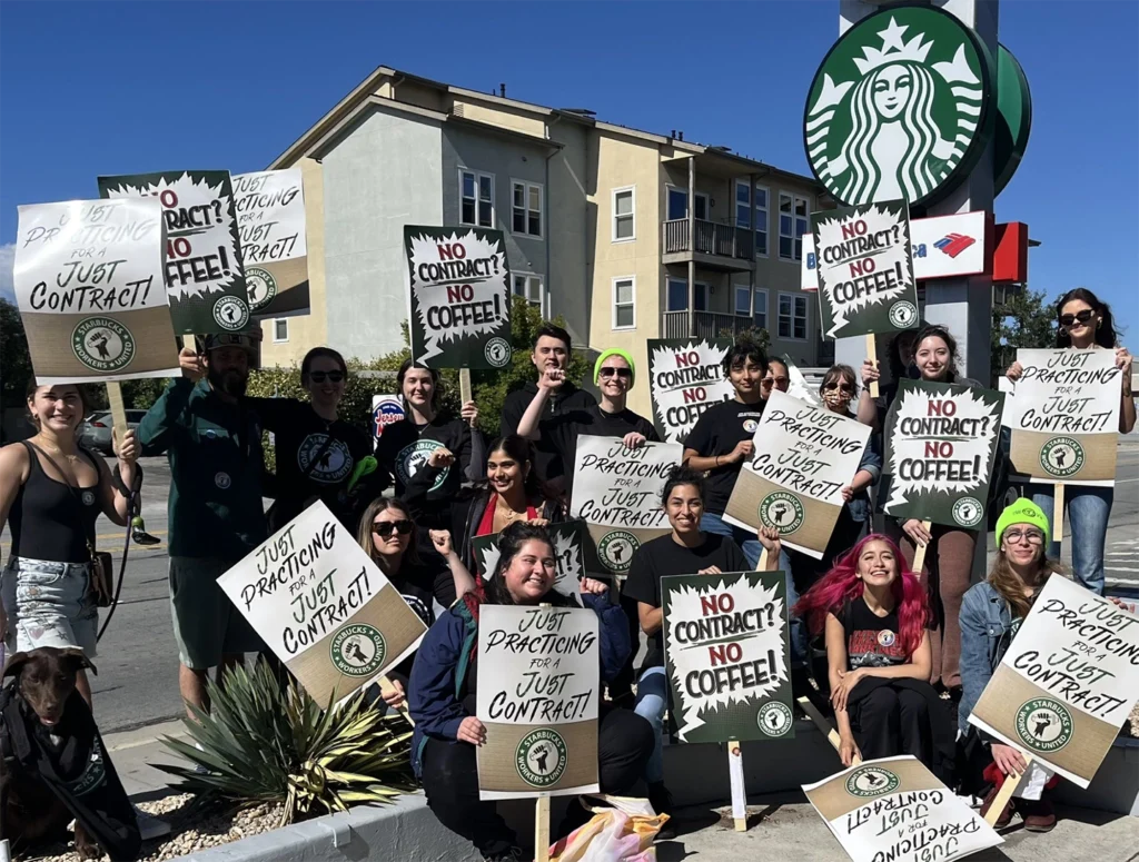 Gruppe von rund zwanzig Personen, die vor einer Starbucks-Filiale protestieren. Sie halten Schilder mit Aufschriften wie „No Contract? No Coffee!“ und „Just Practicing for a Just Contract!“ in den Händen. Die Protestierenden wirken entschlossen und freundlich, viele tragen T-Shirts mit gewerkschaftlichem Logo. Im Hintergrund sind Wohngebäude und das Starbucks-Logo zu sehen, bei sonnigem Wetter.